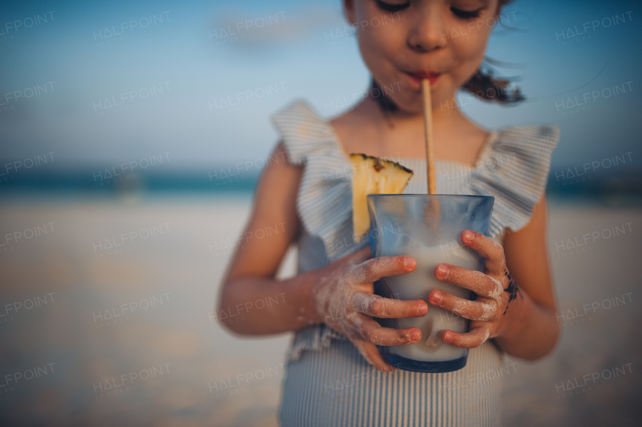 Portrait of a little adorable girl on a sandy beach by the sea. A girl with braids is drinking a non-alcoholic mixed drink. Concept of beach summer vacation with kids.