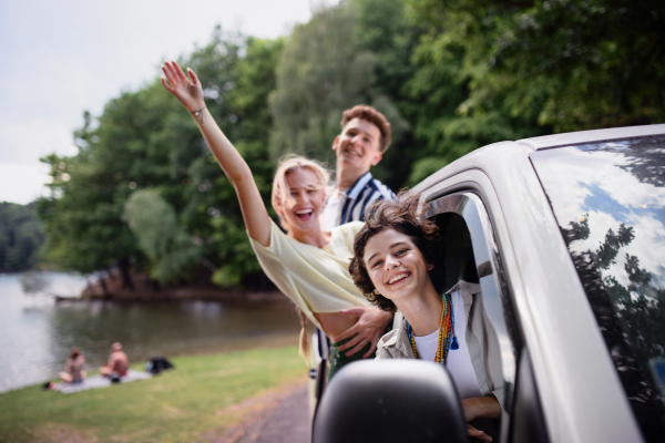 Young friends travelling together by a car, looking through window, waving and smiling - summer vacation, holidays, travel, road trip and people concept.