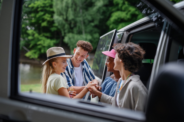 Multiracial young friends travelling together by a car, standing by car and smiling - summer vacation, holidays, travel, road trip and people concept.