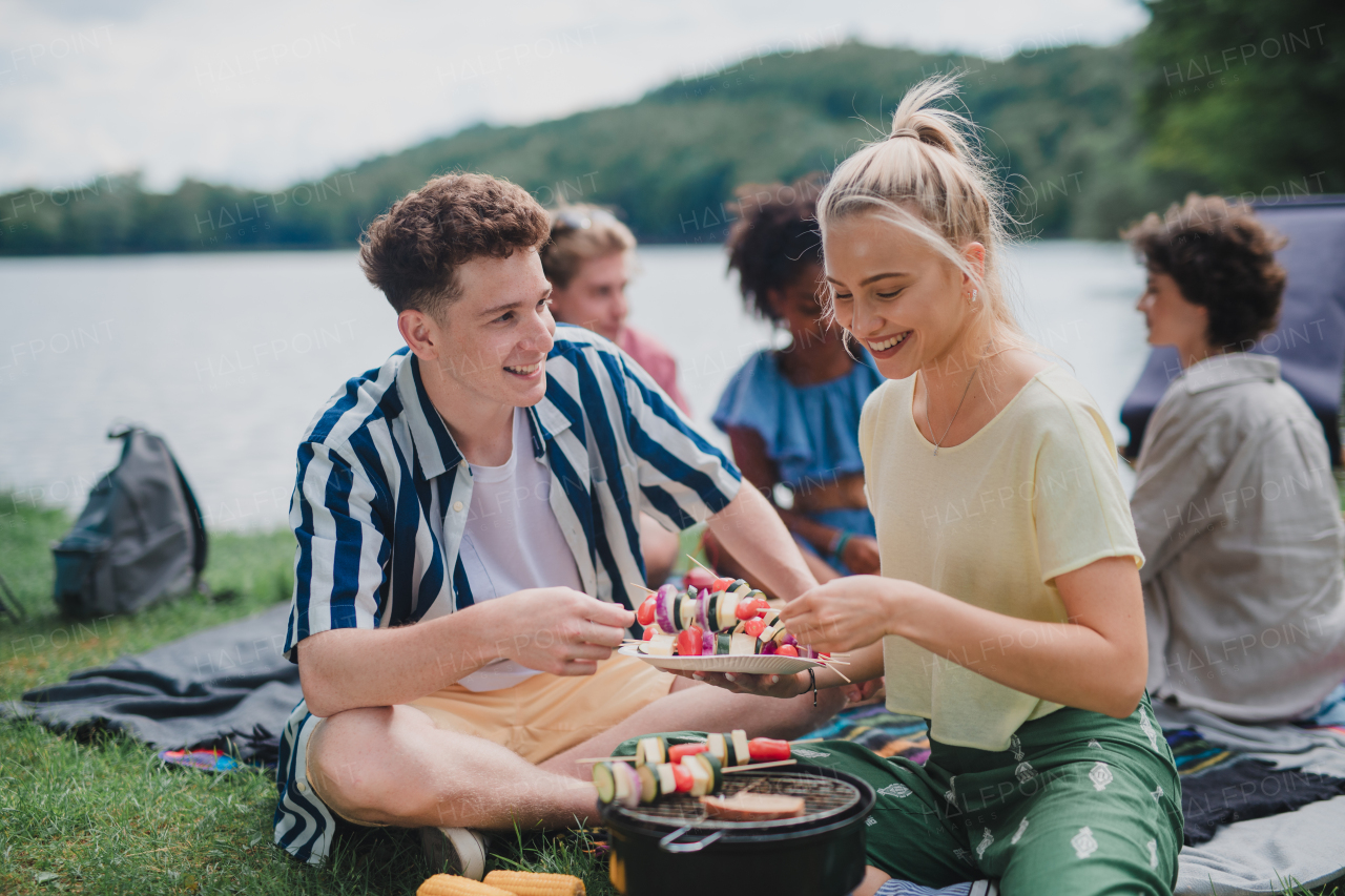 A group of multiracial young friends camping in campsite near lake and and having barbecue, putting skewers o grill.