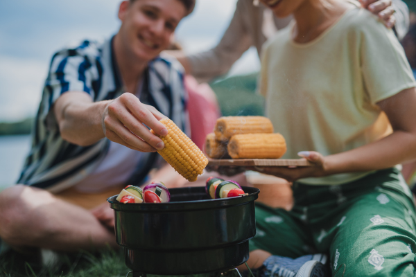 A close-up of young friends putting corn on grill and having barbecue when camping in campground.