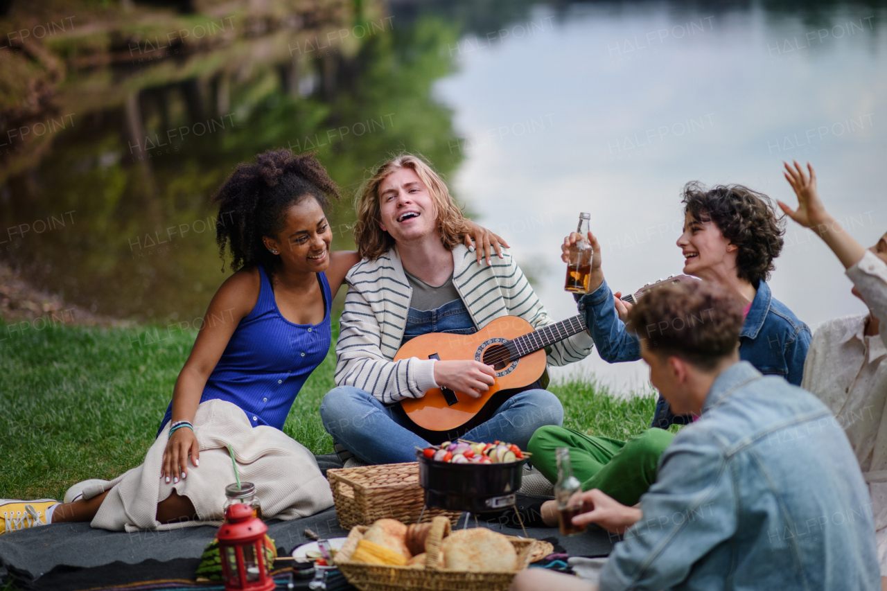 A group of young friends having fun on picnic near a lake, sitting on blanket eating and playing guitar.