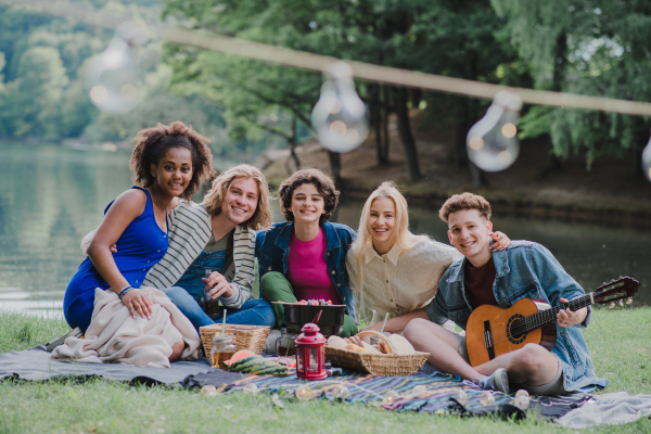 A group of young friends having fun on picnic near a lake, sitting on blanket eating and playing guitar.