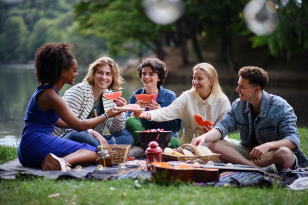 A group of young friends having fun on picnic near a lake, sitting on blanket and eating watermelon.