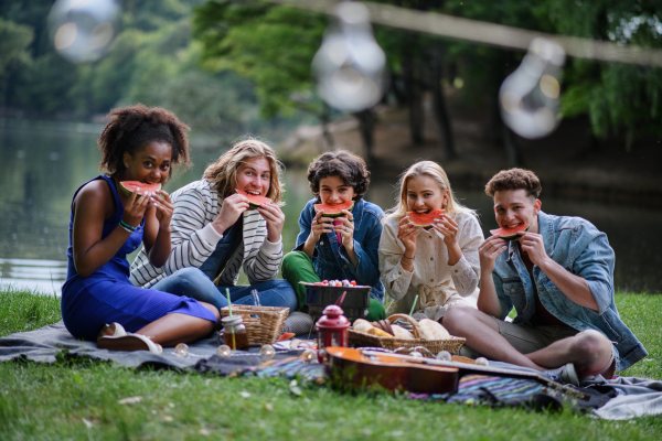 A group of young friends having fun on picnic near a lake, sitting on blanket and eating watermelon.