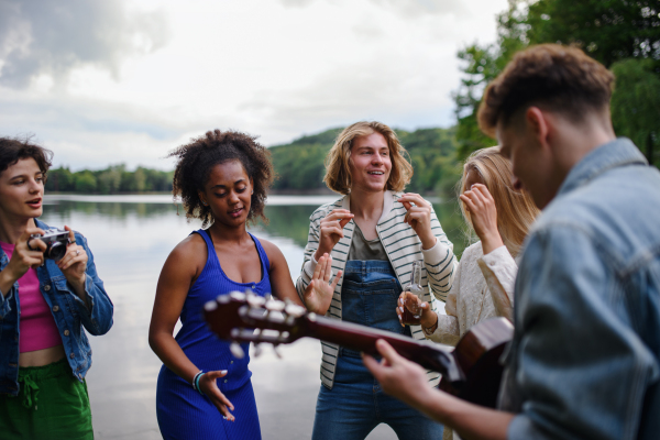 Group of young friends having fun near the lake, laughing and playing guitar.