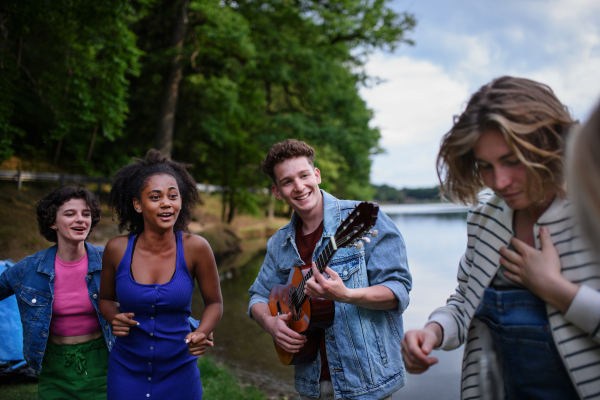 A group of young friends having fun on picnic near a lake, laughing, dancing and playing guitar.