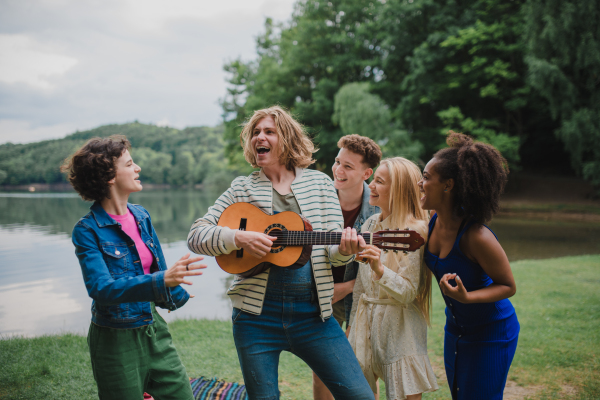 Group of young friends having fun on picnic near a lake, laughing and playing guitar.