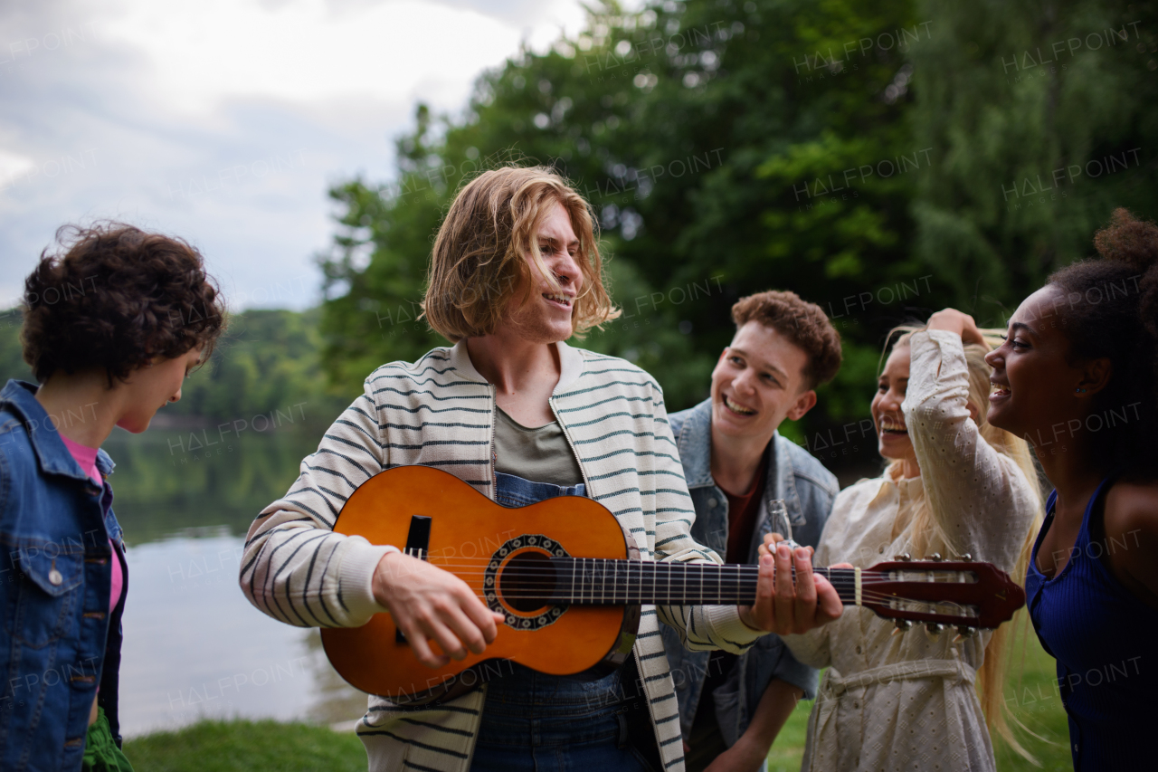 Group of young friends having fun near the lake, laughing and playing guitar.