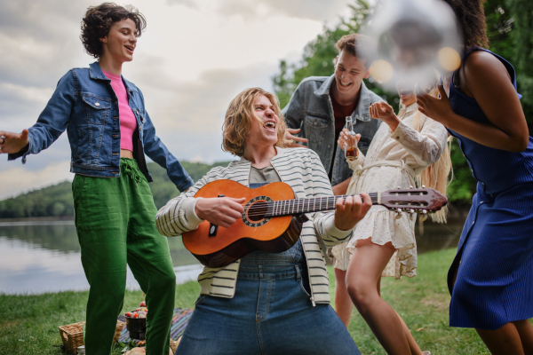 A group of young friends having fun on picnic near a lake, laughing, dancing and playing guitar.