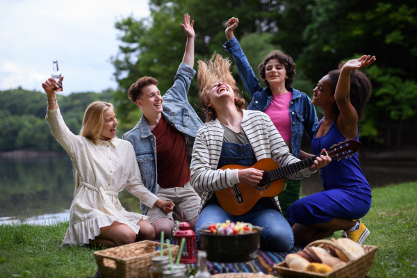 A group of young friends having fun on picnic near a lake, sitting on blanket eating and playing guitar.