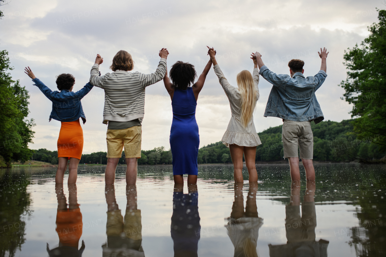 A multiracial group of young friends holding hands and standing in lake in summer. Rear view.