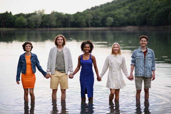 A multiracial group of young friends holding hands and standing in lake in summer.