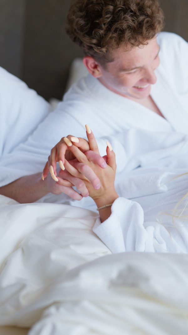 A young couple in bathrobe enjoying together time in wellness hotel room, lying in the bed and hugging.