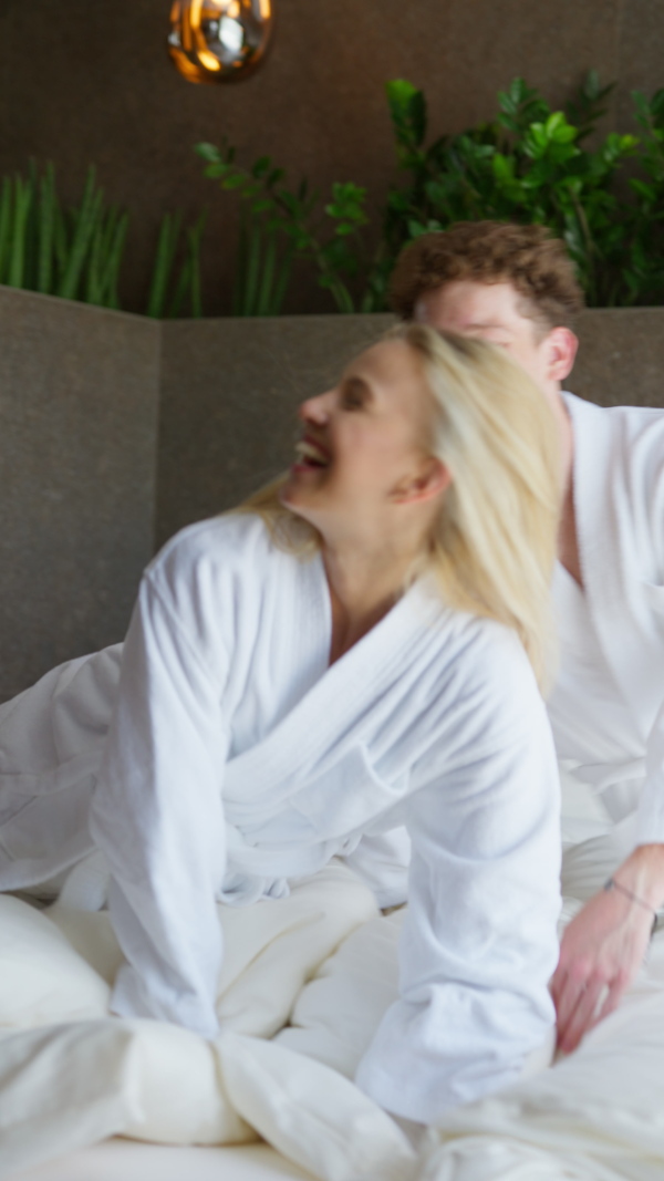 A young couple in bathrobe enjoying together time in wellness hotel room, lying in the bed and hugging.