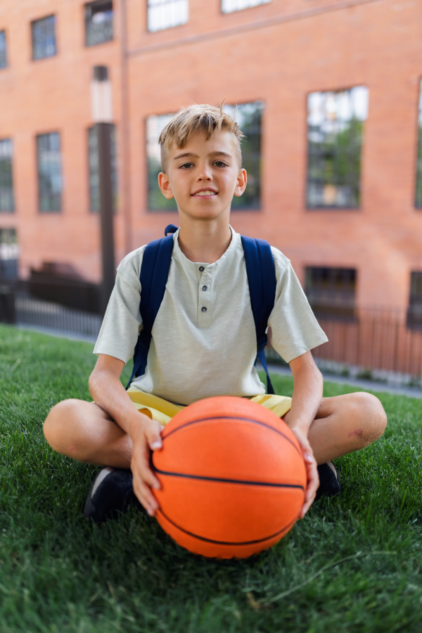 Cheerful caucasian boy sitting in public city park with baskaetball ball and looking at camera.