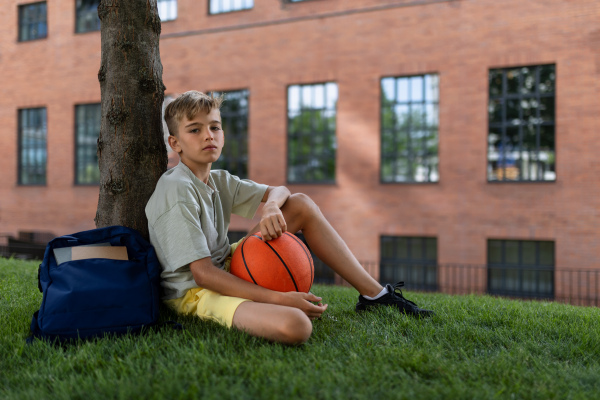 Cheerful caucasian boy sitting in public city park with baskaetball ball and looking at camera.