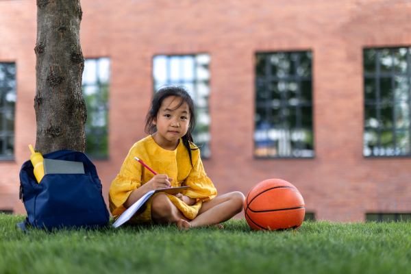 Little asian girl sitting in public park and writing some notes. Summer time.