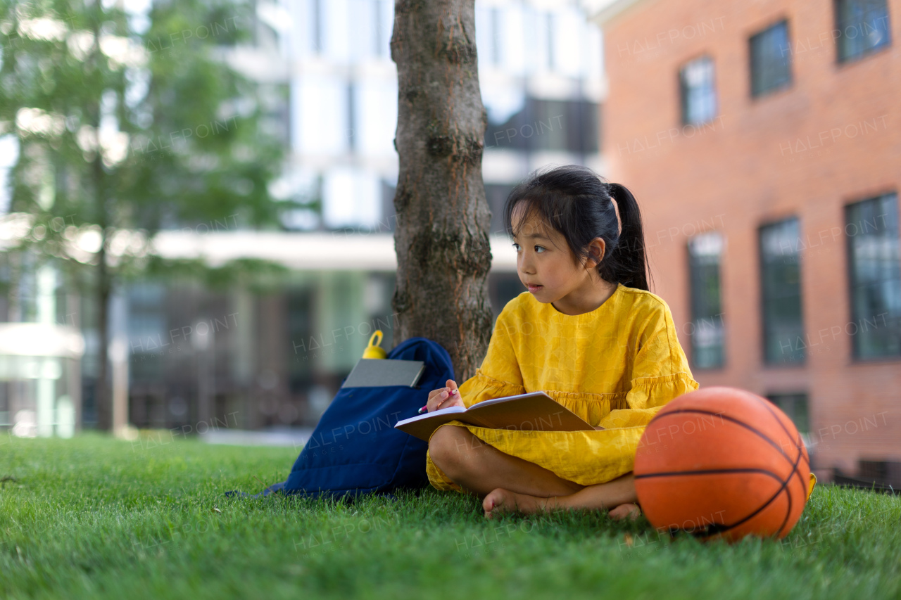 Little asian girl sitting in public park and writing some notes. Summer time.