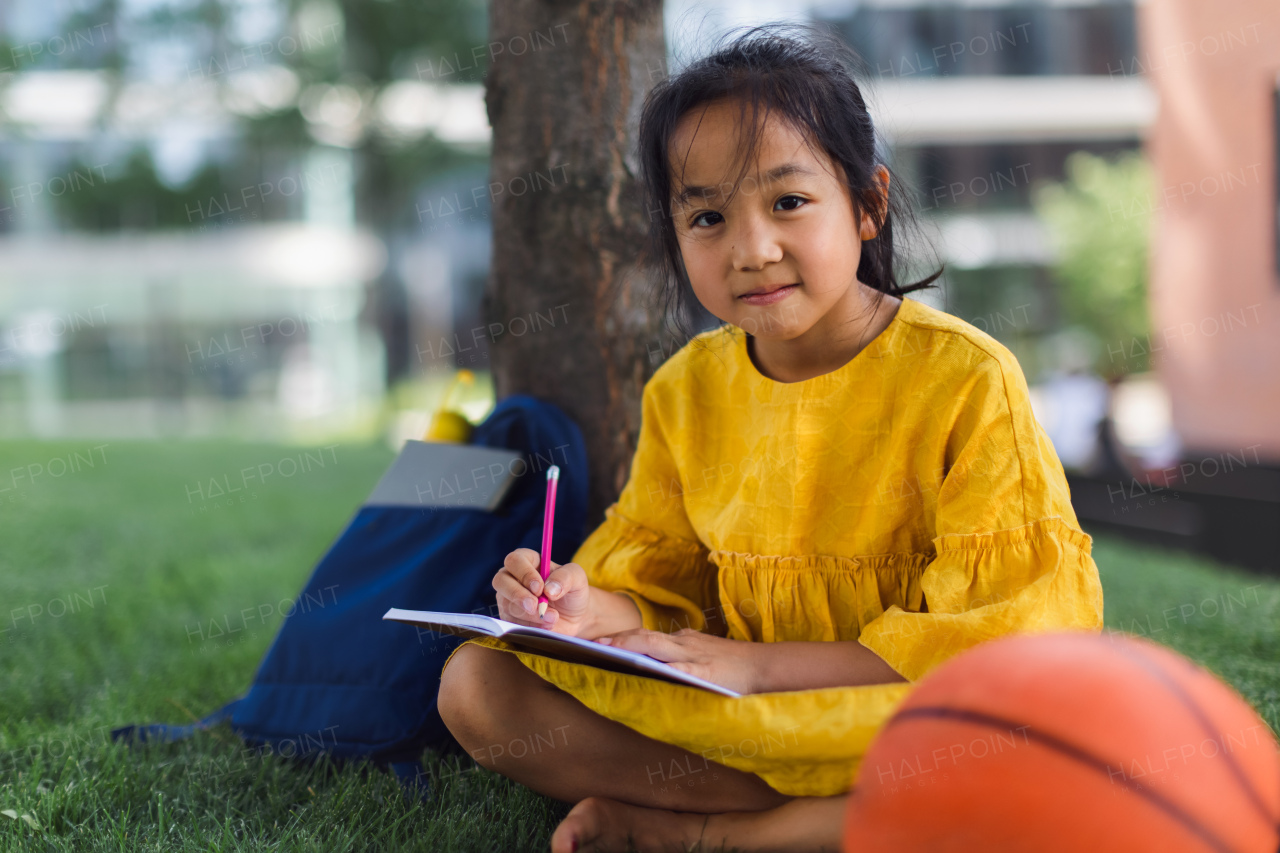 Little asian girl sitting in public park and writing some notes. Summer time.