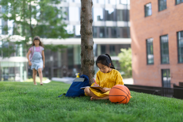 Little asian girl sitting in public park and writing some notes,her friend standing in background. Summer time.