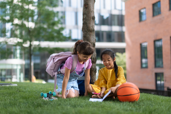 Little asian girl with her friend sitting in public park and writing some notes. Summer time.