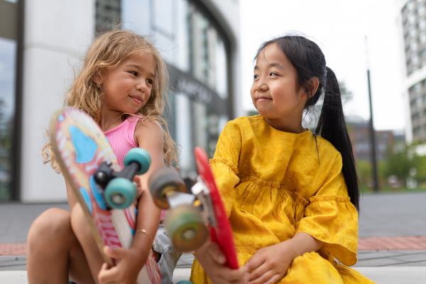 Two girls sitting and holding the skateboards in city street, active kids concept. Low angle view.