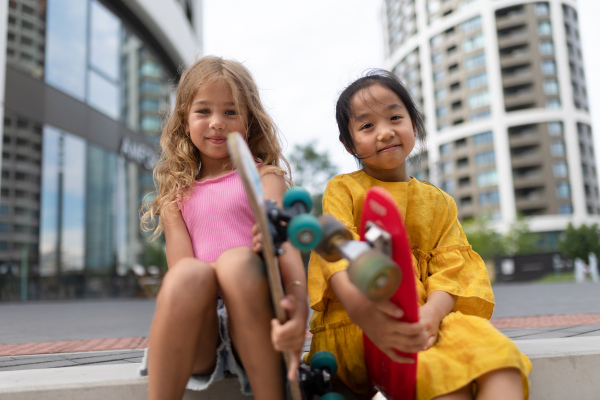 Two girls sitting and holding the skateboards in city street, active kids concept. Low angle view.