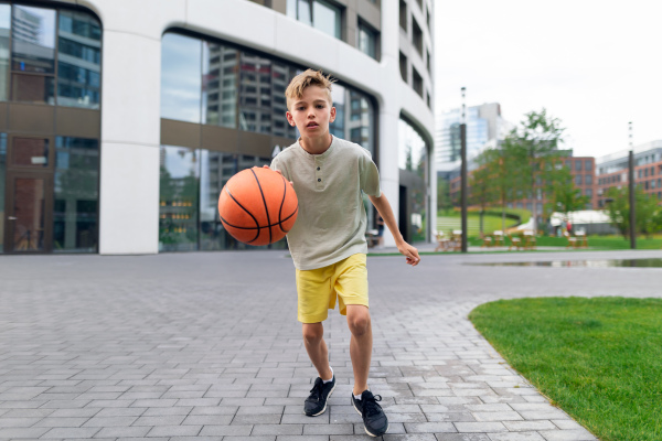 Cheerful caucasian boy driblling with basketball ball in public city park, looking at camera.