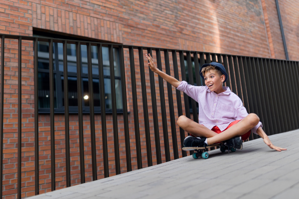 Cheerful caucasian boy sitting in skateboard and going down the hill, looking at camera.