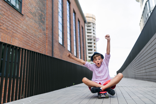 Cheerful caucasian boy sitting in skateboard and going down the hill, looking at camera.