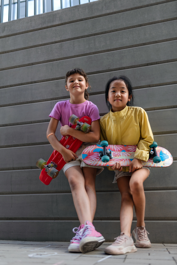 Happy friends posing together in city, standing in front of a concrete wall, holding skateboards and looking at camera.