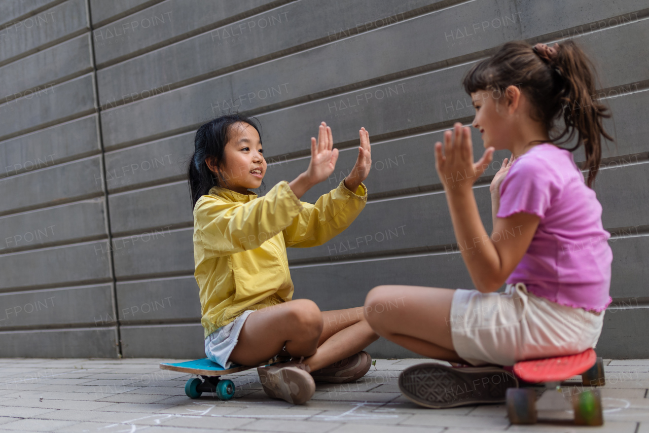 Asian girl with her friend sitting in skateboards in a city street, talking and playing, active lifestyle kids concept.