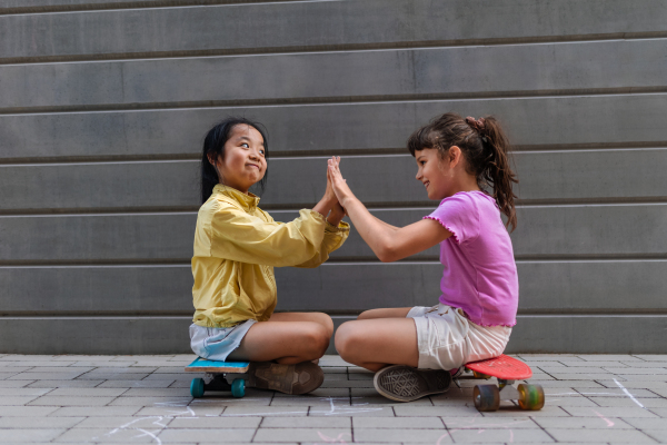 Asian girl with her friend sitting in skateboards in a city street, talking and playing, active lifestyle kids concept.