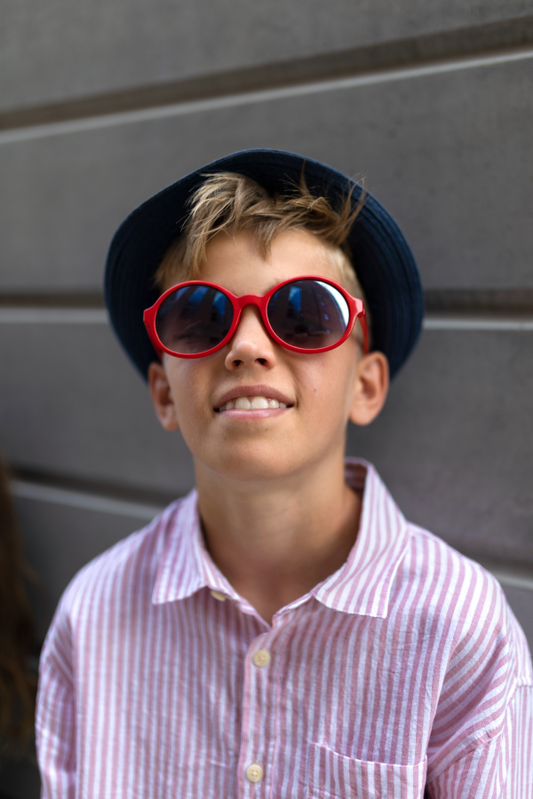 Portrait of caucasian boy in sunglasses and hat standing in urban street in front of a concrete wall.