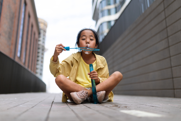 Little girl sitting in a city and blowing bubbles.