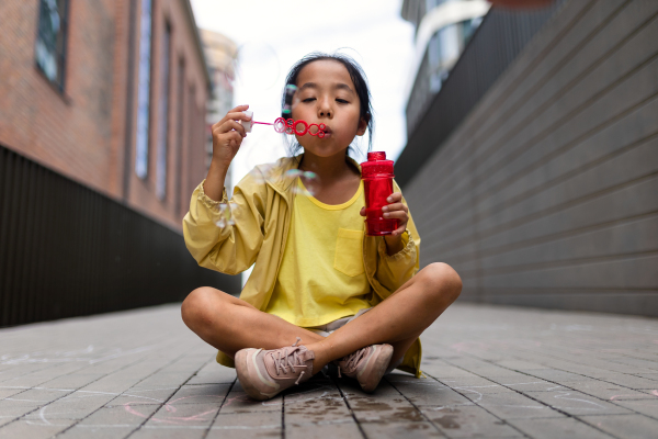 Little girl sitting in a city and blowing bubbles.