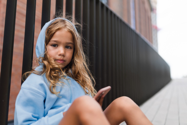 Little blond girl with hood sitting in sidewalk, holding a smartphone, looking at camera.