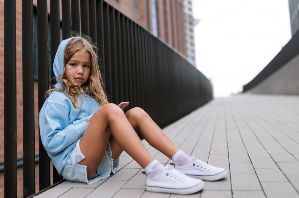 Little blond girl with hood sitting in sidewalk, holding a smartphone, looking at camera.