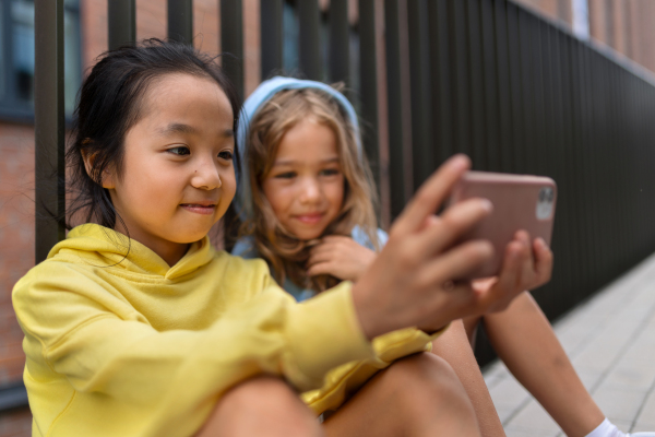 Little girls with hood sitting in sidewalk and scrolling a smartphone.