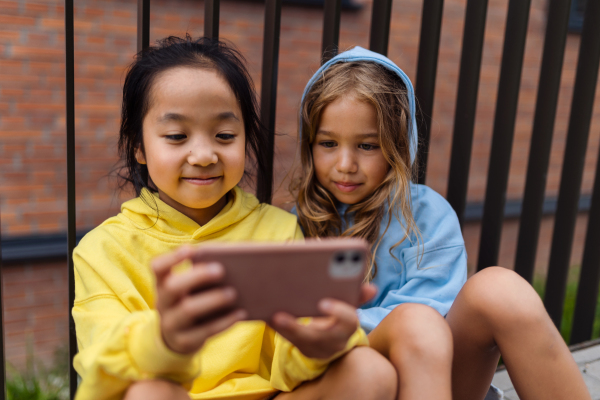 Little girls with hood sitting in sidewalk and scrolling a smartphone.