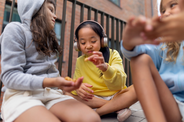 Hapyy three girls friends sitting in sidewalk, talking, listening to music, having a snack and great summer time together. Street style.