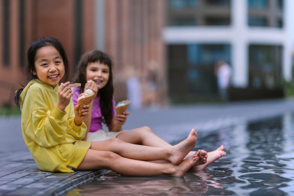 Two girls sitting, having legs in a fountain, enjoying ice cream and summer day.