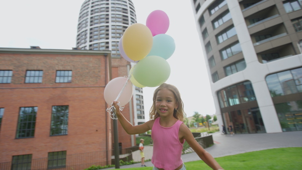 Video of little blond girl running with baloons in city and looking at camera.