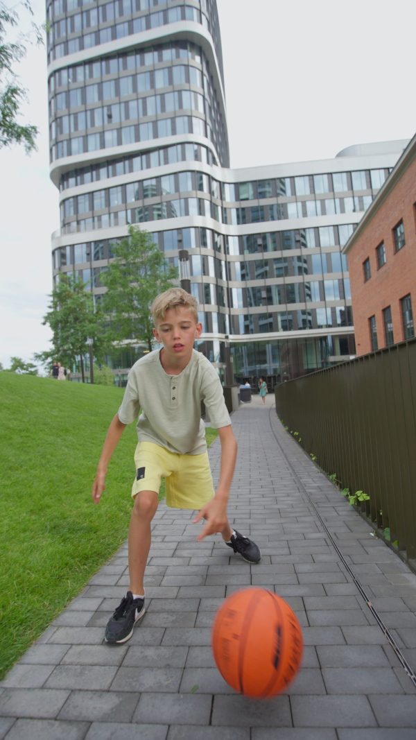 Cheerful caucasian boy driblling with basketball ball in public city park, looking at camera. Vertical footage.