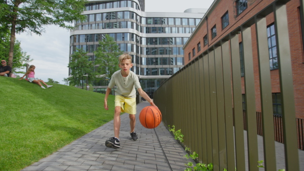 Cheerful caucasian boy driblling with basketball ball in public city park, looking at camera.