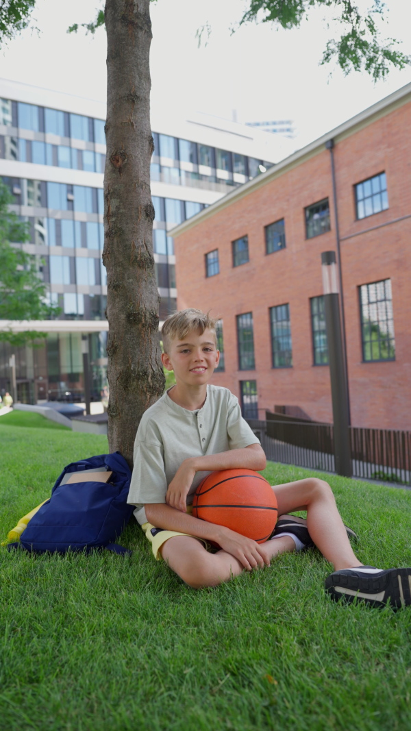 Cheerful caucasian boy sitting in public city park with baskaetball ball and looking at camera. Vertical footage.