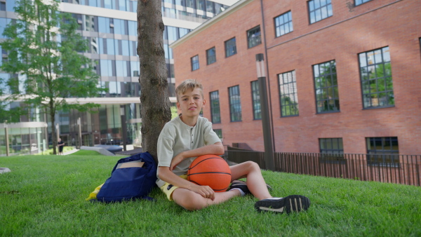 Cheerful caucasian boy sitting in public city park with baskaetball ball and looking at camera.