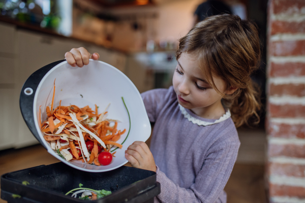 Litte girl putting kitchen waste, peel and leftover vegetables scraps into kitchen compostable waste. Concept of composting kitchen biodegradable waste.