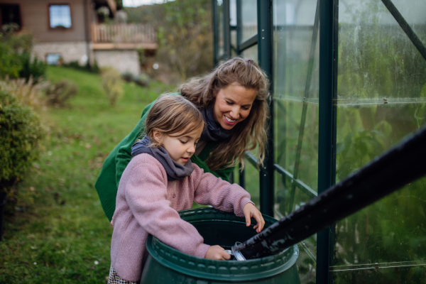 Girl and mother scooping rainwater from barell under downspout, going to water plants in garden. Concept of water conservation in garden.